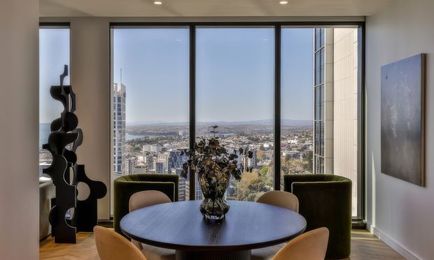 A refined high-rise dining area with a round dark dining table, sculptural centerpiece, upholstered dining chairs, and two deep green lounge chairs positioned in front of expansive floor-to-ceiling windows. The windows provide panoramic urban and distant landscape views. The room has light walls, recessed ceiling lighting, timber flooring, dark window frames, and a large abstract artwork.