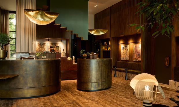 A richly lit, dark-toned hospitality-style interior with two large curved metallic counters, warm timber wall panelling, exposed timber detailing, and decorative pendant lighting. A lounge or reception area extends toward a seating nook with framed artwork and shelving. The space has a dramatic, luxurious atmosphere and a patterned timber floor.