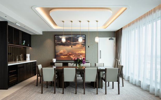 A refined contemporary dining room with a long dark timber dining table surrounded by upholstered chairs. The room features a dark textured feature wall, integrated dark cabinetry and shelving, a large framed artwork, a sculptural recessed ceiling with warm cove lighting, pendant lights, and full-height sheer curtains over expansive glazing.