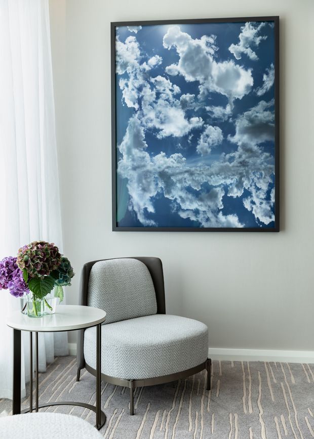 A refined sitting corner with a light grey upholstered accent chair beside a small round white side table holding a vase of flowers. A large framed blue-and-white cloud artwork hangs on a pale grey wall, with sheer curtains at the window and a patterned grey rug underfoot. The room has a calm, tailored contemporary feel.