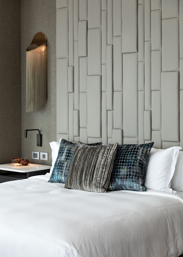 A refined contemporary bedroom featuring a neatly made white bed, layered textured pillows in grey, blue and teal, a sculptural white geometric padded wall feature behind the bed, and a dark textured side wall with a warm metallic wall sconce. A minimalist bedside table and modern electrical outlets complete the sophisticated hotel-like setting.