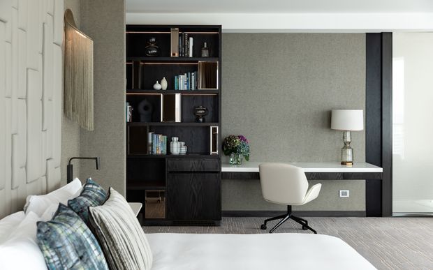 A refined bedroom with a partially visible bed in the foreground, a dark built-in shelving and storage unit, and a compact integrated desk against a textured grey wall. The room features neutral upholstery and bedding, black joinery, a contemporary task chair, layered lighting, and a large window or glazed opening on the right.