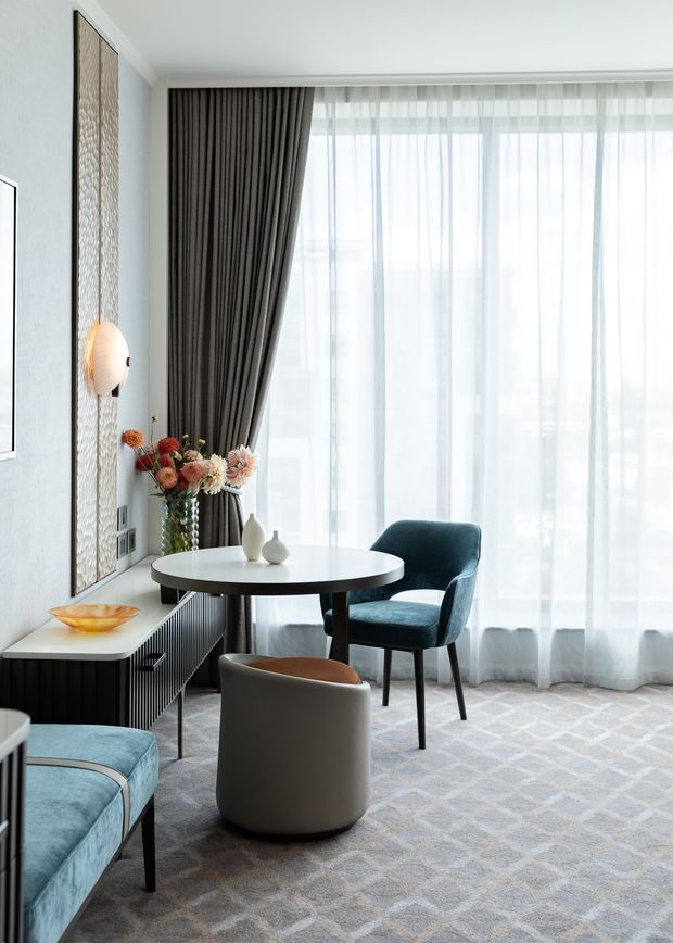 A bright, refined sitting and dining area with floor-to-ceiling sheer curtains and dark drapery, a round black dining table, upholstered dining chairs, a curved lounge chair, and a patterned carpet. The room features a textured decorative wall panel, sculptural wall lighting, a dark built-in sideboard, and a floral arrangement, creating a contemporary hotel-like interior.