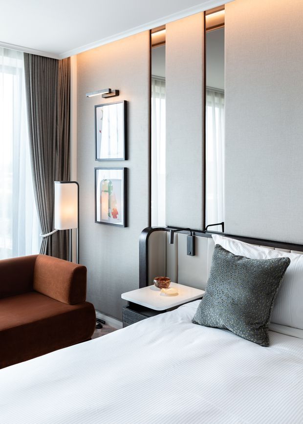 A refined contemporary bedroom with a neatly made bed, upholstered dark headboard, textured grey accent cushion, and an integrated bedside table. The room features full-height mirrored wall panels with warm concealed lighting, textured light-grey wall finishes, framed artwork, sheer curtains with dark drapery, and a brown upholstered lounge chair. The palette is neutral and softly illuminated with warm accent lighting.