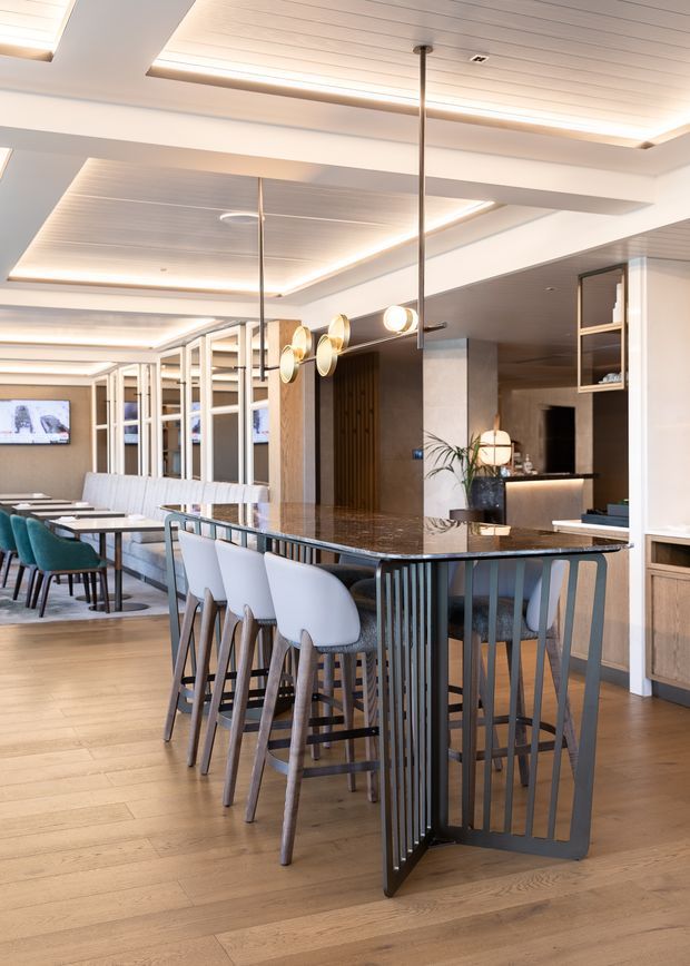 A refined open-plan hospitality or residential entertaining space centred on a long marble-topped bar-height island with dark metal slatted supports and upholstered stools. Behind it is a dining area with a long table, banquette seating and teal chairs, plus a compact illuminated bar or kitchenette. The room features warm timber flooring, light neutral walls, extensive built-in joinery, mirrored partitions, layered recessed and cove lighting, and decorative pendant lights. The overall design is contemporary, polished and warmly lit.