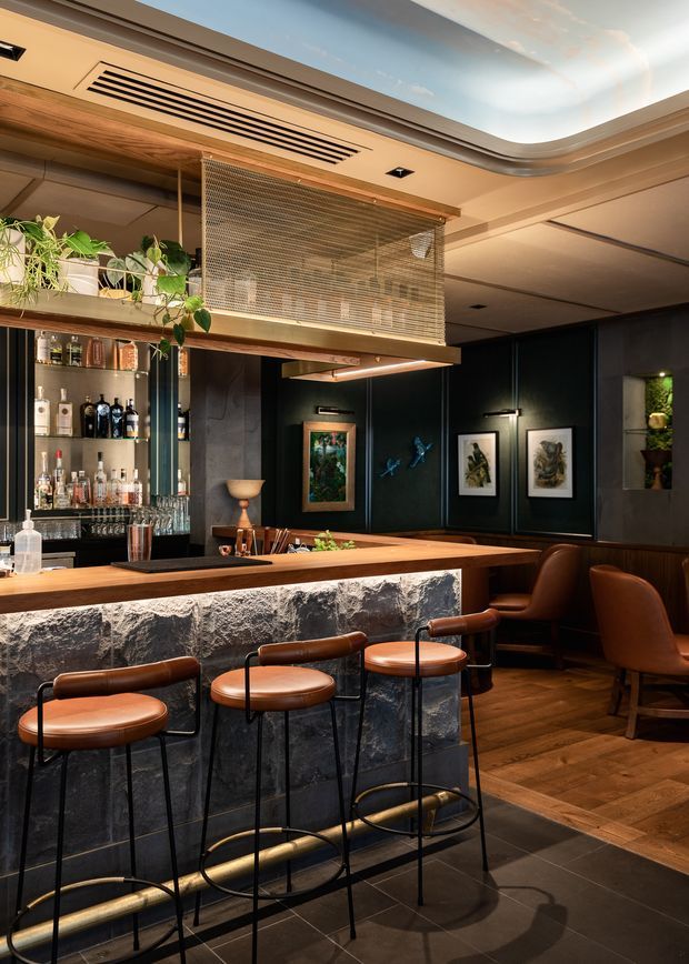 A sophisticated interior bar with a long stone-fronted counter, timber bar top, leather-and-metal stools, dark paneled walls, and extensive back-bar liquor shelving. Warm timber flooring contrasts with dark floor tiles, while integrated lighting, framed artwork, indoor plants, and a decorative mesh mezzanine or overhead screen add layered detail. The space has a moody, refined hospitality-club atmosphere.