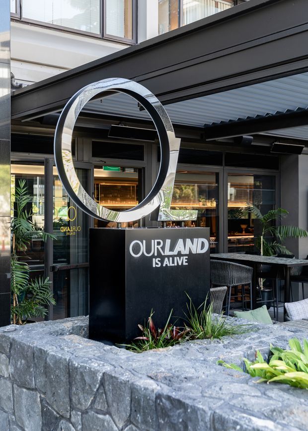 Exterior view of a contemporary commercial hospitality venue with a covered outdoor dining area, dark metal framing, extensive glazing, a large polished circular sign, and a dark stone planter wall with tropical planting. The entrance signage reads “OURLAND IS ALIVE.”