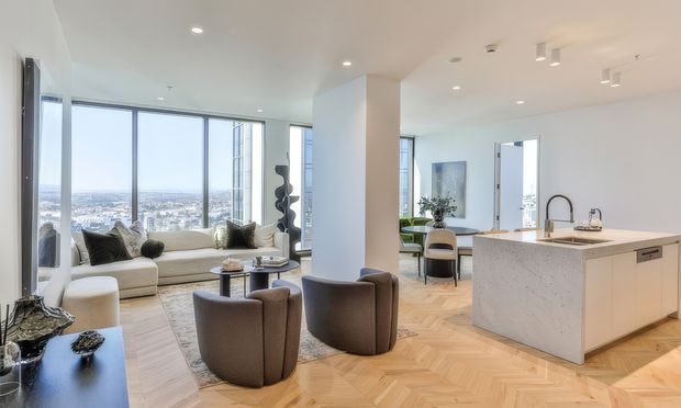 Bright open-plan apartment interior combining a contemporary kitchen, living area, and dining zone. Floor-to-ceiling corner glazing provides expansive city views. The room has pale timber herringbone flooring, white walls and ceiling, a large light upholstered sofa, sculptural rounded lounge chairs, and a substantial white stone kitchen island with an integrated sink and dark tapware. Minimal recessed and track lighting creates an airy, refined atmosphere.