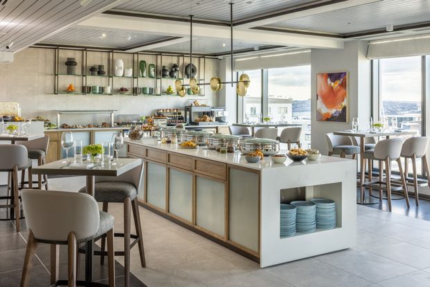A bright, refined open-plan hospitality-style dining and kitchen space with a large central island, integrated shelving, extensive glazing, and city views. The island has light timber-framed cabinetry with pale inset panels, a white benchtop, open dish storage, and multiple buffet-style food displays. Round dining tables and upholstered chairs surround the island, while decorative shelving, pendant lighting, artwork, and a tiled floor complete the contemporary interior.