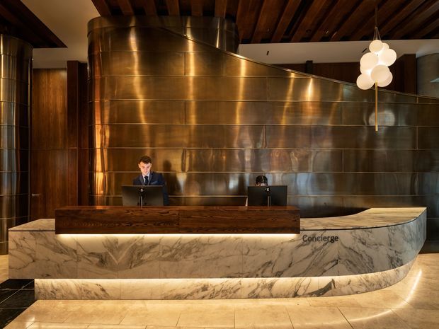 A dramatic hotel or commercial concierge reception area featuring a long curved desk clad in veined marble with warm integrated underlighting and a dark timber reception counter. Behind it is a large sculptural curved wall finished in reflective bronze or copper-toned metal panels, complemented by a dark exposed timber beam ceiling and a clustered globe pendant light. Two reception workstations are visible.