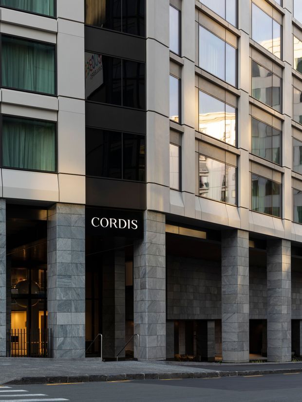 Street-level exterior view of the Cordis hotel building, showing a contemporary multi-storey facade with large rectangular windows, dark glazed vertical bays, pale panel cladding, and substantial grey stone-clad columns and lower walls. A recessed entrance and covered porte-cochère face the street, with the hotel name prominently displayed above the entry.