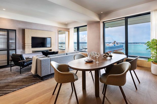 A bright, refined open-plan living and dining area with floor-to-ceiling glazing overlooking turquoise water and a marina. The room features a contemporary dining table with upholstered chairs, a grey sofa, timber flooring, a textured feature wall with wall-mounted television, black-framed glazing, and layered recessed lighting.
