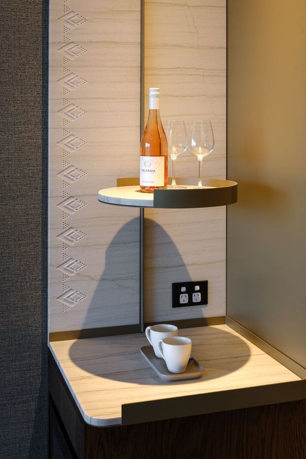 A compact hospitality-style beverage station featuring a stone-look or marble-look tiled wall with a geometric decorative inlay, dark wood cabinetry, and a pale stone-look benchtop with rounded edges. A small circular black-and-light shelf holds a wine bottle and two glasses, while two white cups sit on a tray below. Warm directional lighting highlights the refined built-in joinery and wall detailing.