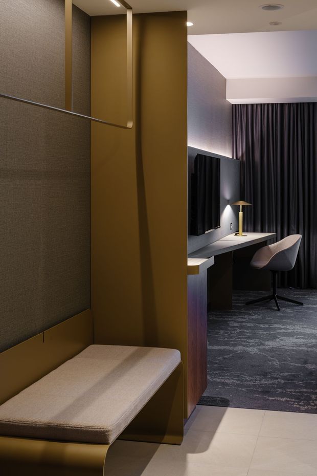 A sophisticated compact interior combining a built-in upholstered bench or daybed in the foreground with a dark, refined home office beyond. The office features a wall-mounted black television or display, a long integrated desk, a sculptural upholstered task chair, a slim brass-gold table lamp, heavy charcoal curtains, and textured dark carpet. The space is divided by a tall ochre-brown architectural partition with integrated lighting and textured wall surfaces. The overall atmosphere is moody, luxurious, and highly tailored, with warm accent lighting and contrasting pale floor tiles.