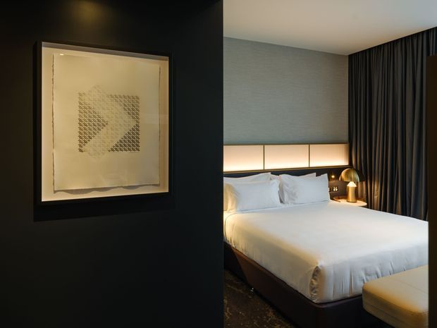A moody, refined contemporary bedroom viewed through a dark entry wall. The room features a neatly made bed with white bedding, a padded grey upholstered headboard, integrated warm bedside lighting, a metallic table lamp, full-height dark curtains, and a cream upholstered bench. A framed abstract artwork is mounted on the black wall in the foreground.