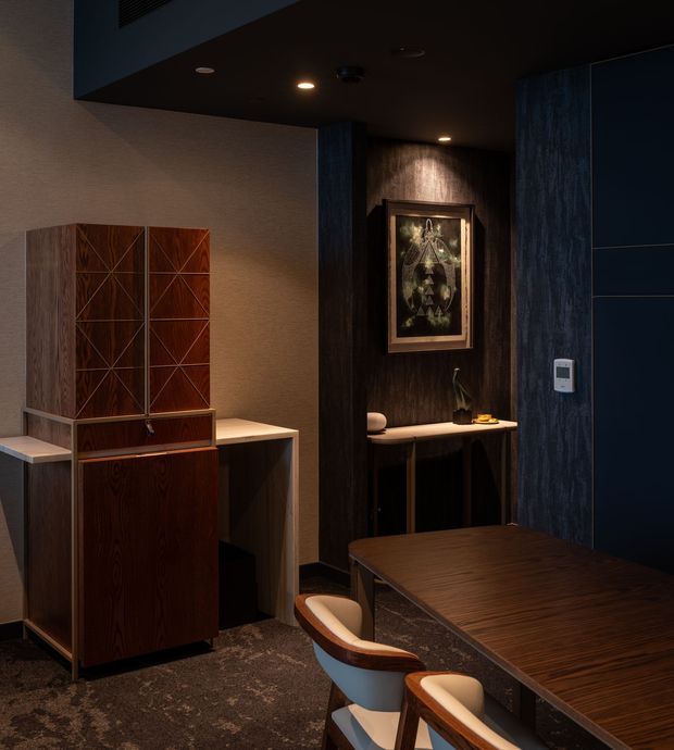 A dark, richly detailed dining and entertaining interior with a large dark timber dining table and sculptural upholstered chairs in the foreground. A walnut-toned built-in cabinet and desk-like console sit against a textured beige wall, while a timber-lined alcove with artwork and a narrow console adds depth. The room features dark ceilings, blue-black wall panels, warm recessed lighting, and patterned dark carpet, creating an intimate, sophisticated atmosphere.
