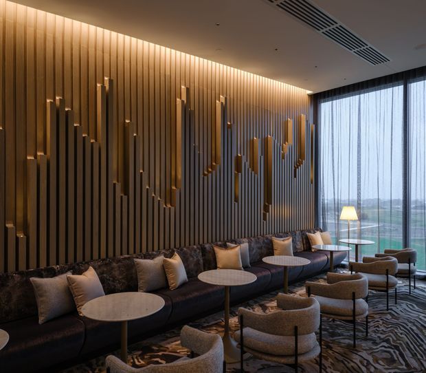 A sophisticated lounge or hotel-style reception area with a long dark sectional sofa, clustered round café tables, upholstered armchairs, and floor-to-ceiling sheer curtains overlooking a green landscape. The dominant feature is a warm backlit timber slat wall with sculptural vertical relief detailing. A patterned carpet, contemporary lighting, and muted brown, beige, charcoal, and gold tones create a refined, intimate atmosphere.