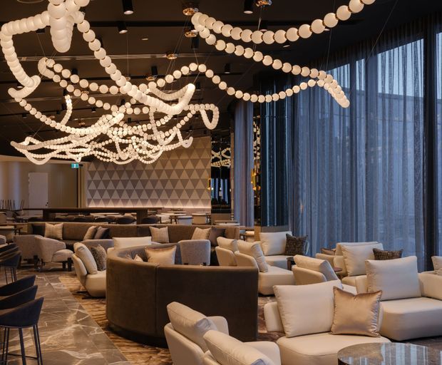 A spacious, upscale lounge or hospitality interior with numerous cream and white upholstered sofas and armchairs arranged around low tables. Dramatic strands of spherical pendant lights sweep across a dark ceiling, while floor-to-ceiling sheer curtains line the glazed exterior wall. A geometric triangular feature wall and patterned stone flooring add visual interest, creating a refined, warm, contemporary atmosphere.