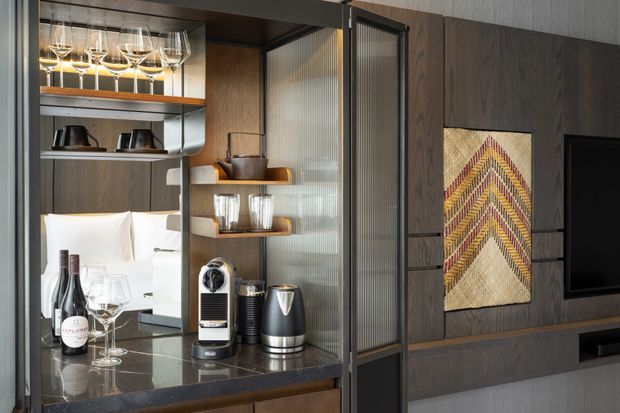 Close-up of a refined built-in beverage and coffee station integrated into dark timber wall cabinetry. The joinery includes glassware storage, open shelves, a stone-look benchtop, coffee equipment, wine, and ribbed translucent glass sliding panels. A geometric woven artwork and television are visible on the adjoining dark timber media wall.
