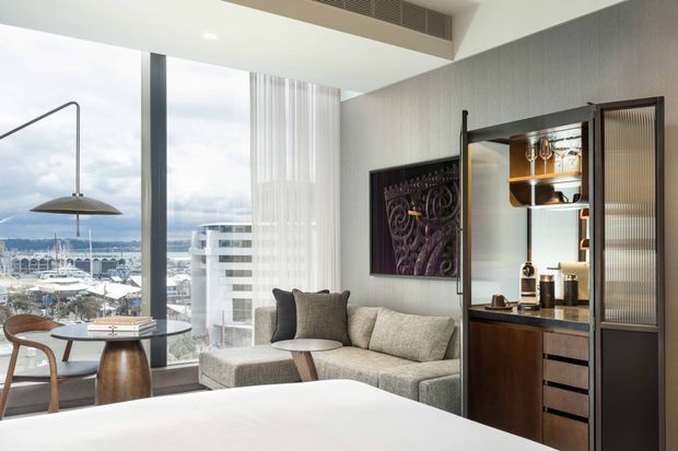 A refined modern hotel-style bedroom or studio suite with a large bed in the foreground, a textured neutral sofa, compact round dining table and chairs, full-height glazing overlooking a waterfront cityscape, and a dark timber built-in bar or kitchenette cabinet with illuminated shelving. The space uses clean lines, contemporary furniture, a neutral palette, and integrated ceiling ventilation.
