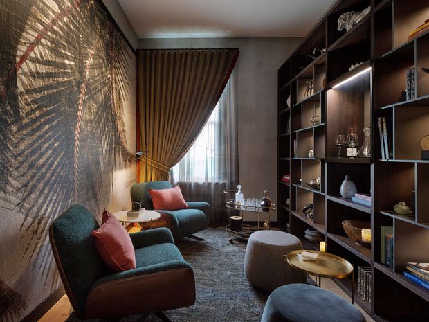 A refined, moody living room featuring deep teal upholstered lounge chairs and ottomans, rust-red cushions, dark timber built-in shelving, layered ambient lighting, patterned wall covering, and full-height drapery framing a window. Decorative objects, books, and a small drinks table create a sophisticated sitting and reading area.
