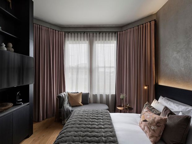 A moody, refined bedroom with a large bed, upholstered grey quilt, layered pillows, a compact lounge chair, full-height mauve curtains, sheer window coverings, dark built-in cabinetry, and a textured grey feature wall behind the bed. Warm concealed lighting and timber flooring create a cosy atmosphere.