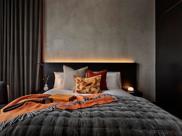 Moody contemporary bedroom with a low upholstered bed, charcoal quilted bedding, layered neutral and rust-toned pillows, and a warm concealed light strip behind the dark headboard. The room features a textured grey plaster wall, full-height dark curtains, black built-in cabinetry, bedside lighting, and a decorative throw across the bed.