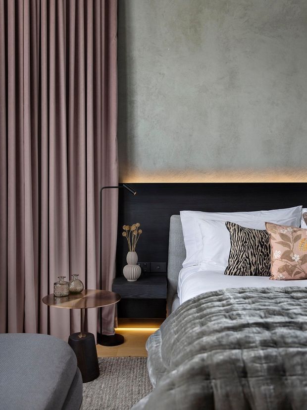 A sophisticated bedroom interior featuring a low-profile bed with a dark timber headboard, layered white bedding, patterned accent cushions, and a textured grey throw. Full-height mauve curtains frame the left side, while a softly textured grey wall is illuminated by concealed warm lighting above the headboard. A small round side table, slim black floor lamp, bedside lighting, and subtle decorative objects complete the refined, moody setting.