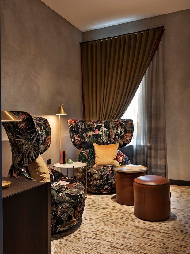 A richly layered sitting area with sculptural floral-patterned armchairs, a small round side table, leather ottomans, and a warm brass floor lamp. Textured taupe walls, a large pleated brown curtain with sheer glazing behind it, and a striped neutral rug create an intimate, theatrical atmosphere.