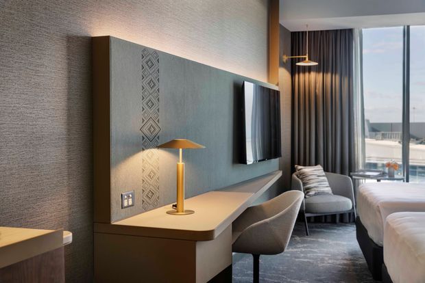 A refined contemporary hotel-style bedroom with two beds, a wall-mounted television, and a built-in desk and dressing area. The room features a textured grey feature wall with geometric decorative detailing, warm wood-toned joinery, integrated lighting, pendant lights, upholstered desk chairs, full-height dark curtains, and large glazing overlooking an exterior view.