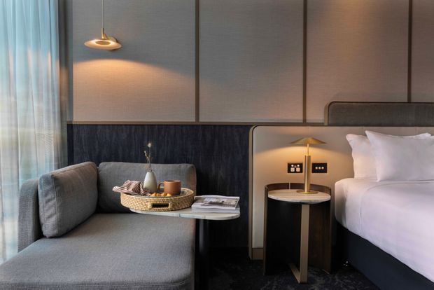 A refined contemporary bedroom corner featuring a grey upholstered window seat or chaise, a bedside table with a slim brass lamp, and a partially visible bed with white linens. The room has dark textured wall panelling, warm ambient lighting, floor-to-ceiling sheer curtains, and a muted neutral palette with brass accents.