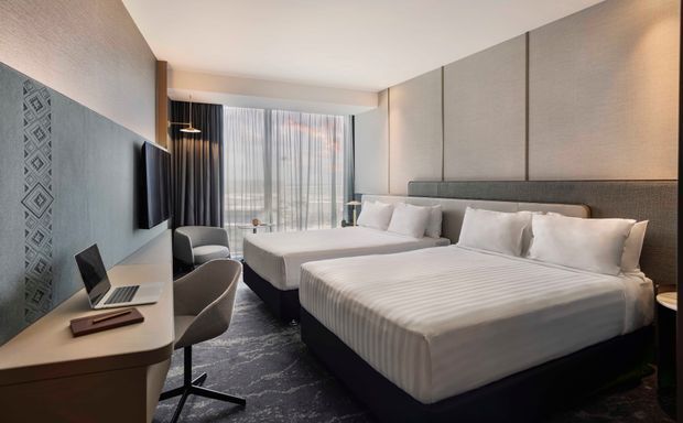 A contemporary hotel-style twin bedroom with two neatly made beds, upholstered headboards, a wall-mounted television, compact work desk, task chair, armchair, and floor-to-ceiling sheer curtains. The room uses a restrained palette of grey, beige, white, and dark wood, with textured wall panels and patterned carpet.