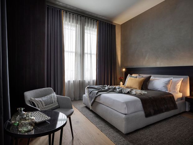 A moody, refined bedroom with a large upholstered bed, layered neutral bedding, dark full-height curtains, a textured grey feature wall, and warm timber flooring. A sculptural black round side table with decorative objects and a grey lounge chair occupy the foreground, while soft daylight filters through sheer curtains.
