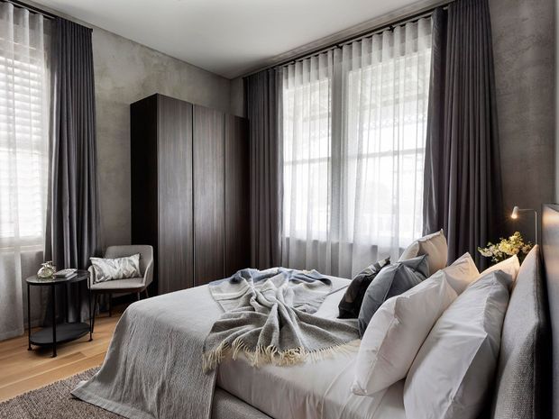 A refined bedroom with a large upholstered bed, layered white bedding and a grey fringed throw. Floor-to-ceiling sheer and dark charcoal curtains frame tall windows, while a dark timber wardrobe, textured grey walls, timber flooring, and a small side table create a moody, contemporary interior.