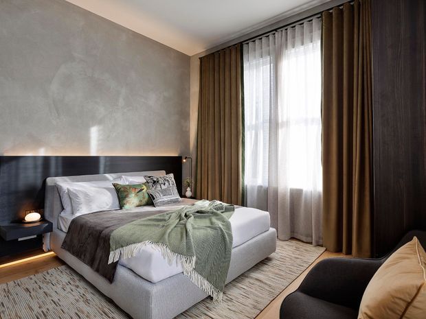 A sophisticated contemporary bedroom with a low upholstered bed, dark wood headboard and integrated bedside shelf, textured taupe feature wall, full-height layered curtains, and dark timber wardrobe cabinetry. Neutral bedding, a sage green throw, patterned cushions, and a warm layered rug create a refined, cosy atmosphere.