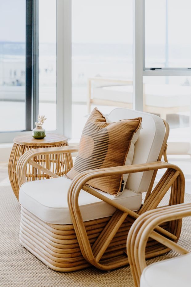 Bright, airy interior seating vignette with a rattan lounge chair and matching ottoman, white cushions, and a striped tan throw pillow. A small round rattan side table with a plant sits beside the chair, while large white-framed windows provide abundant natural light and a soft coastal outlook. The room has a textured neutral carpet and warm woven timber/rattan detailing.