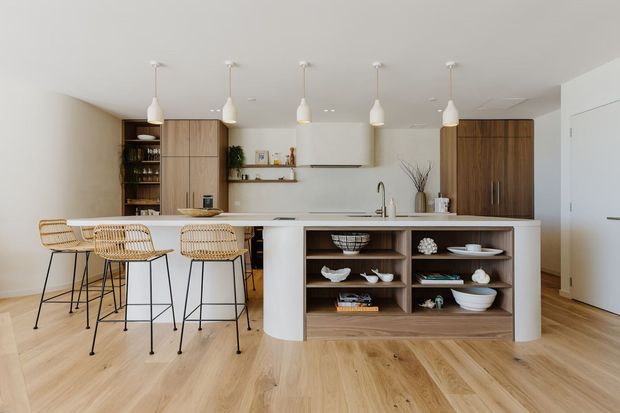 Bright contemporary kitchen with a large white waterfall island and integrated open shelving, surrounded by warm timber cabinetry and light timber flooring. Four woven rattan bar stools sit at the island, with pendant lights above, open shelves and kitchen appliances visible along the rear wall. The room has a clean, warm, refined aesthetic with generous open floor area.