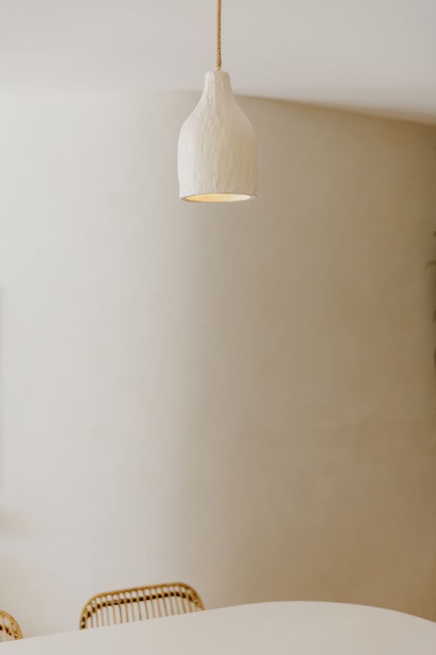 Minimal, softly lit dining area featuring a sculptural white pendant light suspended from the ceiling, warm neutral plaster walls, and the edge of a light dining table with woven rattan chairs visible at the bottom of the frame.
