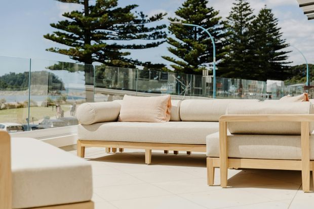 A bright outdoor terrace overlooking a waterfront, furnished with a large cream upholstered sofa, matching armchair and low ottoman. Clear glass balustrades frame views of the water and tall coastal trees, while pale tiled flooring and light timber furniture create a relaxed resort-like setting.