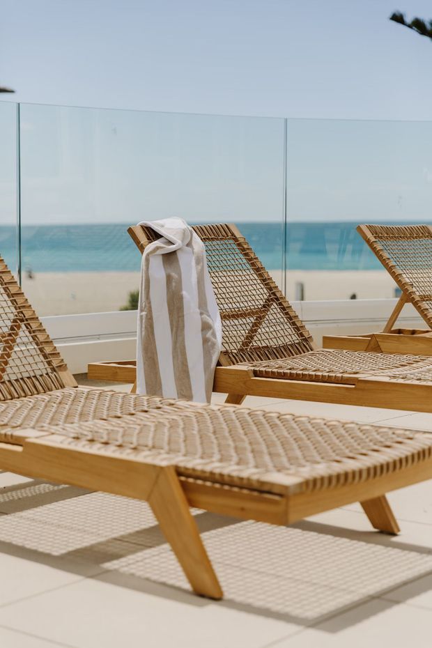 A sunlit seaside terrace furnished with several woven timber sun loungers, one draped with a light striped towel. A clear glass balustrade frames views of a sandy beach and blue ocean beyond. The terrace has pale tiled flooring and a relaxed resort-like atmosphere.