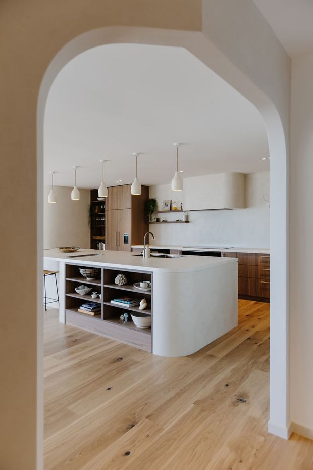A warm, refined contemporary kitchen viewed through a large rounded archway. The room features a substantial curved island with an integrated sink and open display shelving, light timber flooring, dark timber cabinetry, pale plaster walls, a slim linear rangehood, and multiple sculptural pendant lights. The kitchen has a calm, minimalist material palette with softly rounded architectural forms.