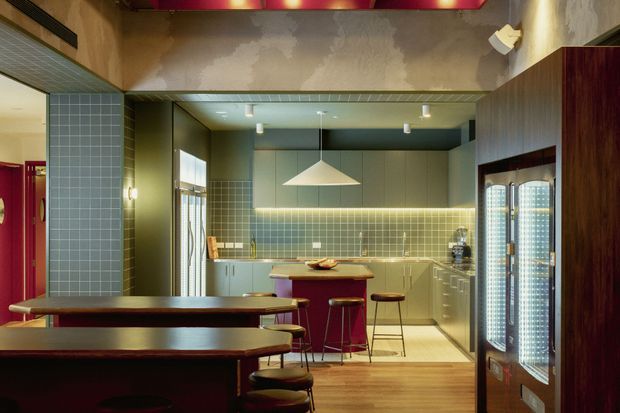 A stylish open-plan kitchen and dining area with a central island, integrated cabinetry, tiled splashback, pendant light, timber flooring, and built-in booth seating. The space features dark timber joinery, muted green-grey tiles, warm under-cabinet lighting, and a glass-fronted beverage refrigerator, creating a moody hospitality-inspired atmosphere.