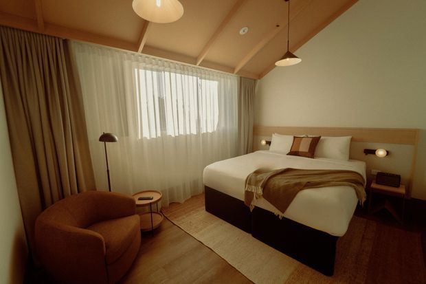 Warm, softly lit bedroom with a large upholstered bed, integrated timber-toned headboard, neutral bedding, sheer curtains, layered drapes, a rust-coloured lounge chair, small side tables, pendant lights, and timber flooring. The room has a cozy mid-century-inspired hotel or residential character with a pitched, exposed-beam ceiling.