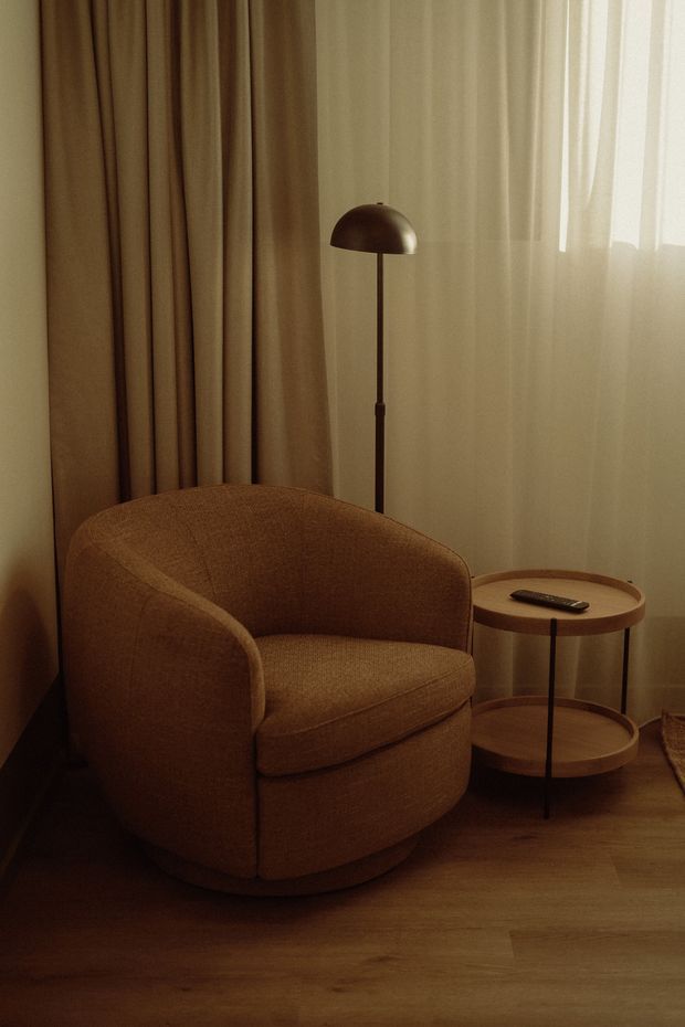 A warmly lit, softly styled corner of a living room featuring a rounded upholstered lounge chair in brown fabric, a slim dark floor lamp with a dome shade, and a small two-tier round timber side table. Floor-to-ceiling curtains and sheer window coverings create a calm, intimate atmosphere, with timber flooring beneath.