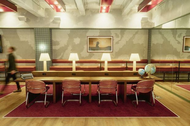 A retro-inspired communal workspace or lounge with a long timber worktable, four red upholstered metal-framed chairs, desk lamps, a red area rug, and a built-in red banquette along the back wall. The room features exposed concrete ceiling beams, distressed textured wall finishes, large mirrors, globe decor, framed artwork, and warm ambient lighting, creating a distinctive mid-century and industrial atmosphere.