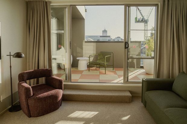 A warm, mid-century-influenced living room with a broad sliding glass opening onto a small balcony or terrace. The room has beige carpet, full-height taupe curtains, a textured burgundy armchair, an olive-green sofa, and a vintage-style floor lamp. Outside, the terrace has terracotta and cream checkerboard tiles, a green metal chair, a small white table, and views of nearby urban buildings and construction cranes.