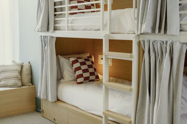 Built-in bunk bed arrangement in a compact bedroom, featuring two stacked sleeping berths with white timber ladder rails, warm timber cabinetry, integrated storage drawers, and grey curtains for privacy. The lower bunk has white bedding and red-and-white checked cushions, while a small upholstered bench or seating area sits beside the window.
