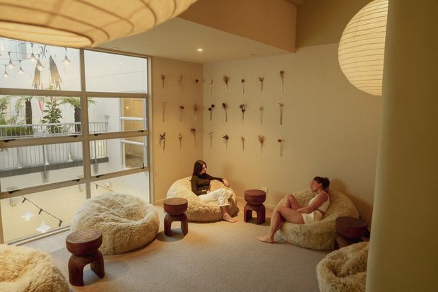 Warm, softly lit lounge or relaxation space with low, rounded furry seating, small dark timber side tables, and delicate dried botanical wall arrangements. Large windows overlook an interior void or adjacent space, while oversized paper lanterns create a calm, intimate atmosphere.