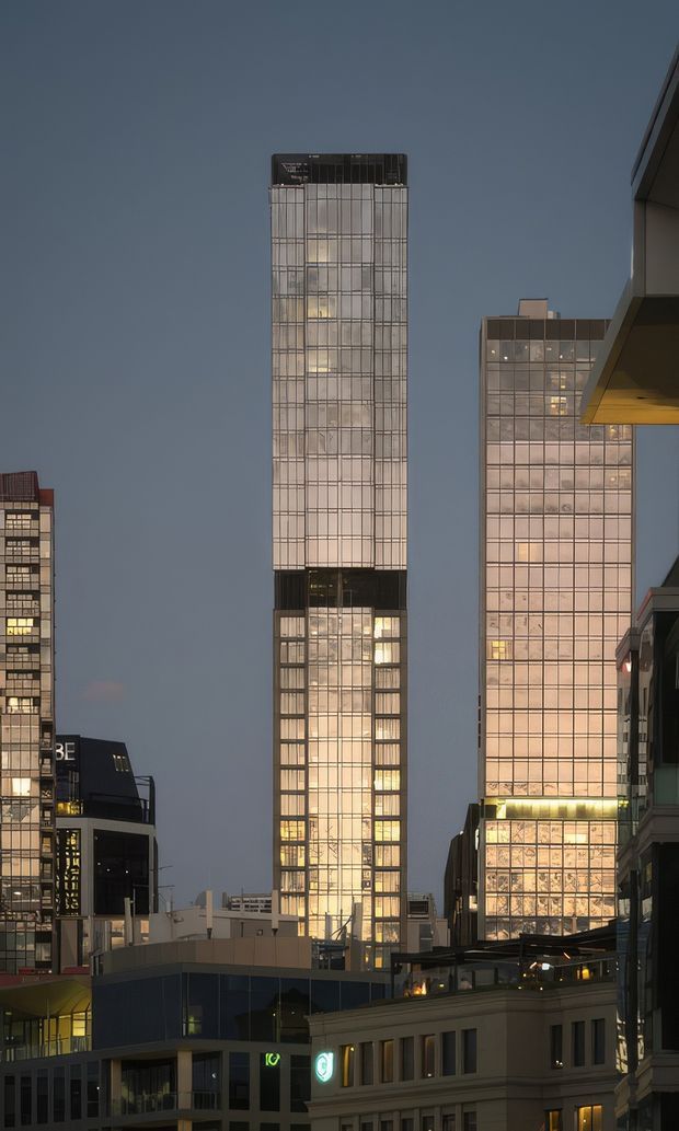 A dusk-time urban cityscape focused on several illuminated high-rise commercial buildings. The central tower has a tall, narrow rectangular form with a gridded glass facade and a darker structural band near its middle, while the adjacent tower has a broad illuminated curtain-wall grid. Lower-rise commercial buildings and illuminated signage frame the foreground.