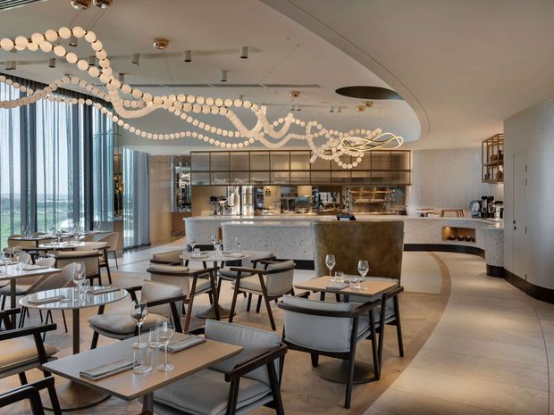 A refined contemporary restaurant interior with a spacious dining area, numerous neatly set tables, a curved central bar and service counter, open shelving, and floor-to-ceiling glazing overlooking a distant landscape. Decorative sculptural pendant lighting made from looping strands of spherical elements spans the ceiling. The palette combines warm beige flooring and surfaces with light upholstered seating, dark timber or metal frames, and brass-toned accents.